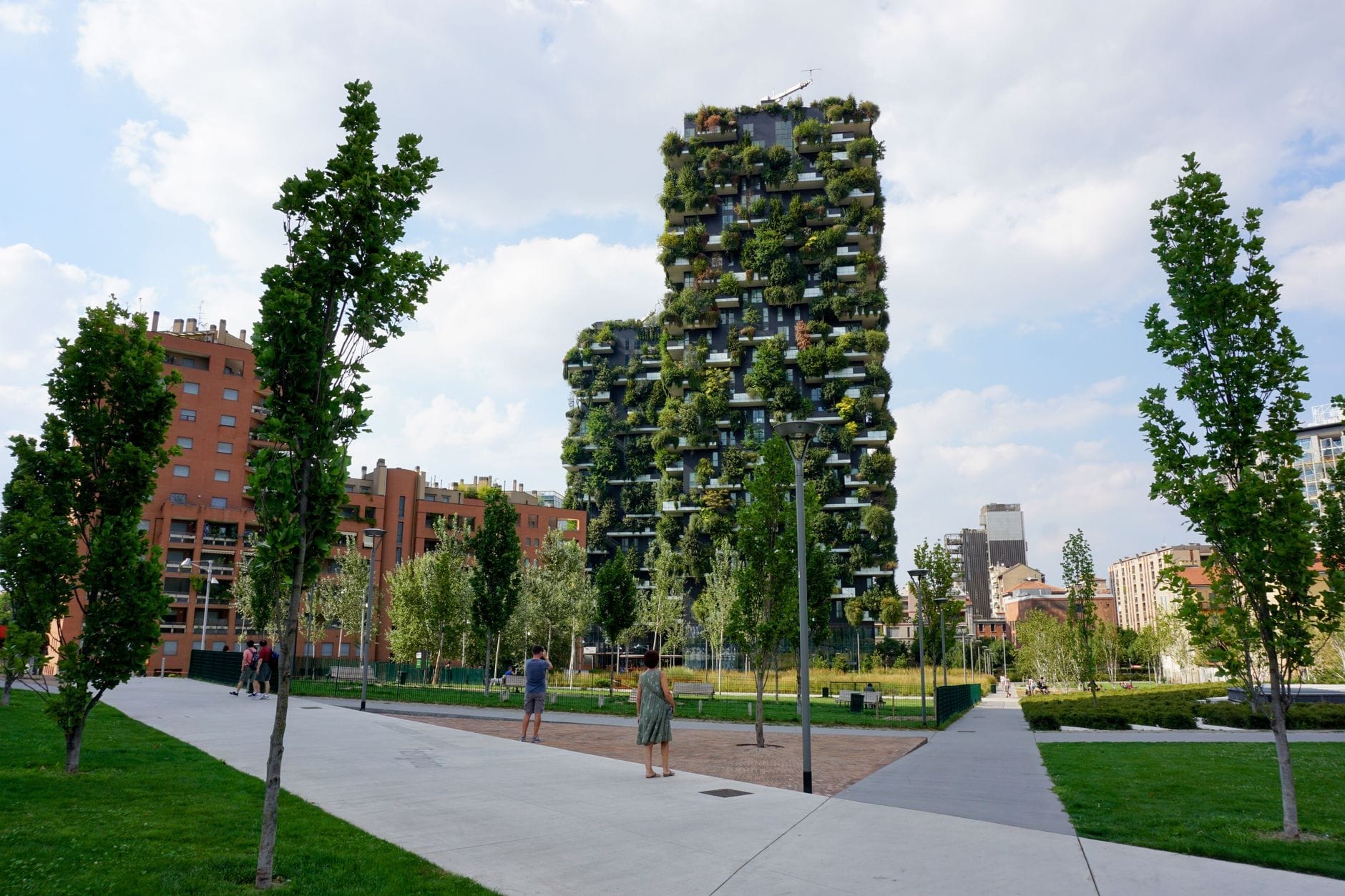 Le rigogliose torri verdi del Bosco Verticale si innalzano alle spalle di un parco urbano con vialetti alberati, giovani piante e persone che passeggiano sotto un cielo luminoso e parzialmente nuvoloso.