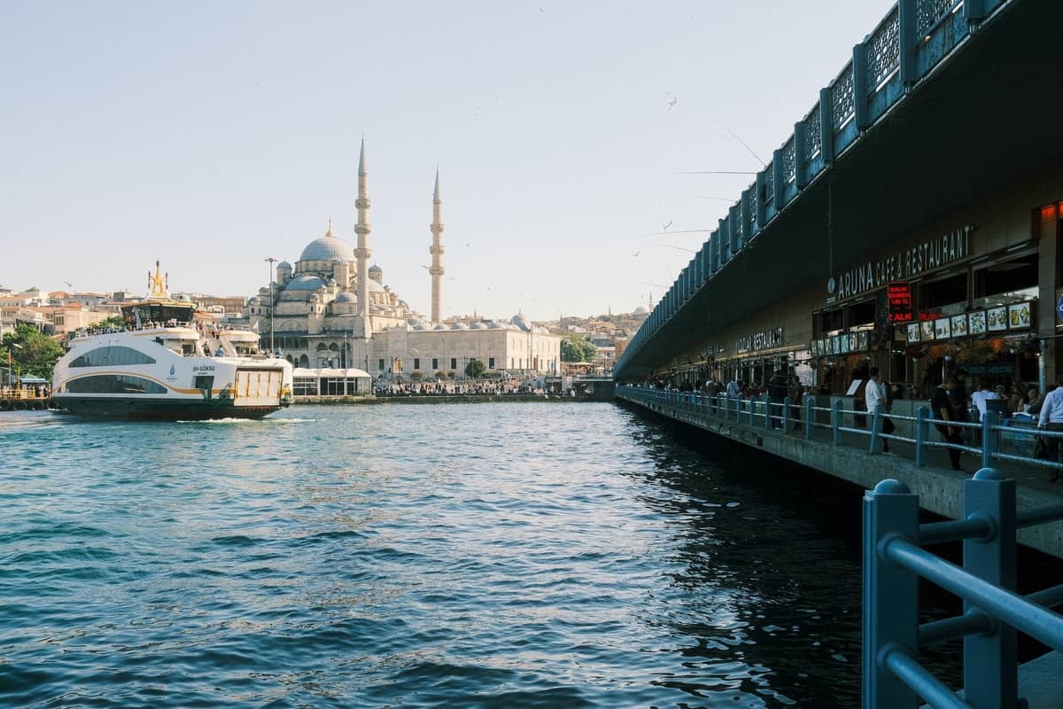 Widok na molo Eminönü i Most Galata nad Bosforem z promem i widocznym Nowym Meczetem, ludzie spacerujący wzdłuż nabrzeża.