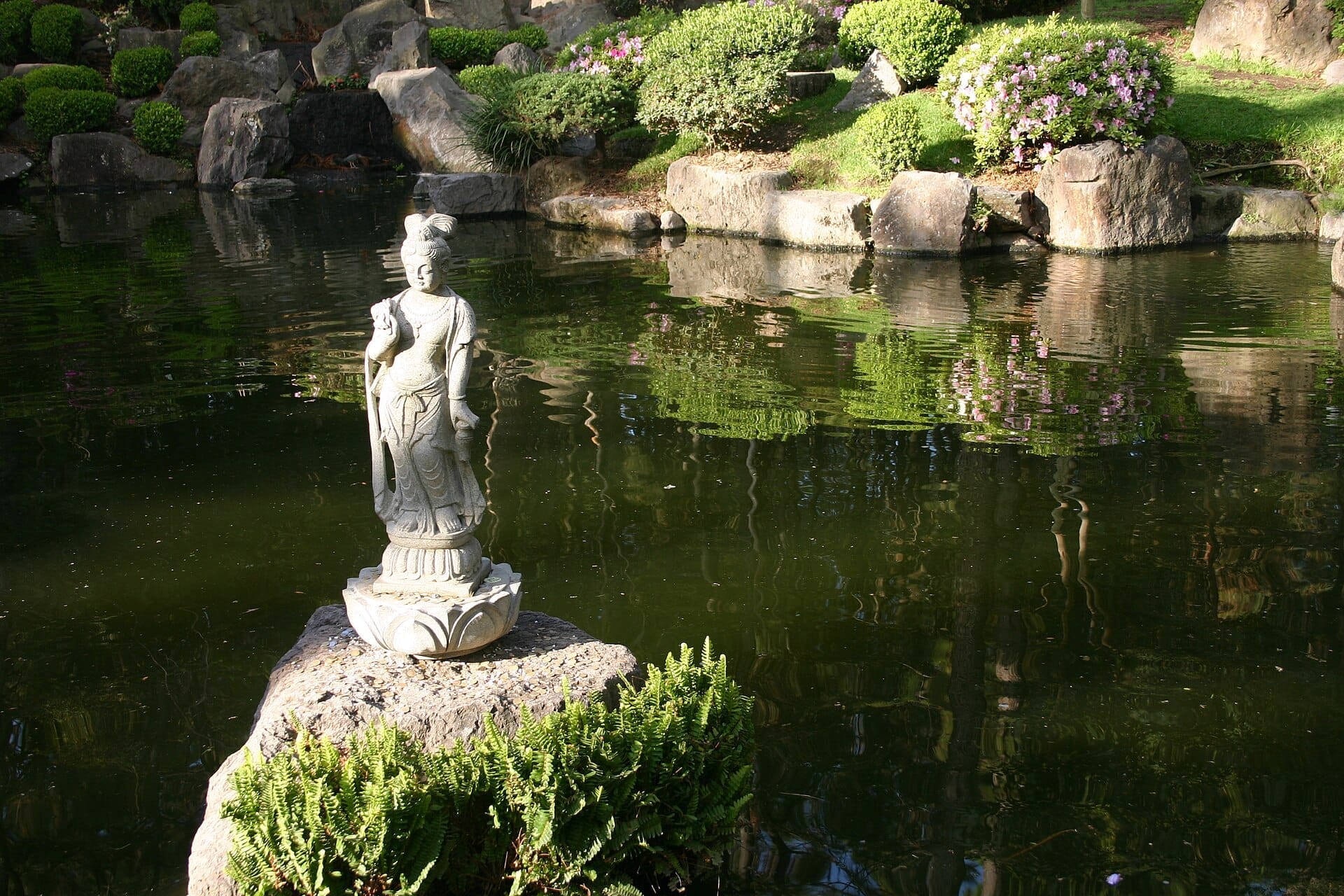 Statua di pietra su una roccia accanto a uno stagno tranquillo, rigogliosi cespugli verdi e fiori in fiore nel Jardín Japonés del Bosque Los Colomos.