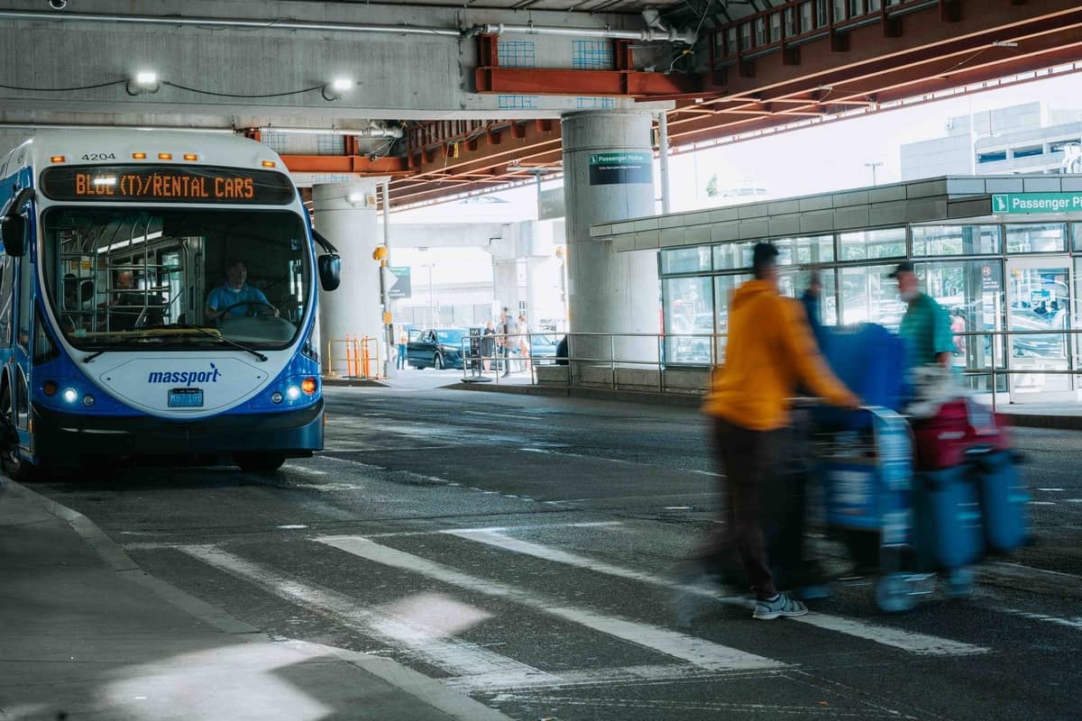Ein Massport-Shuttlebus mit der Aufschrift 'Rental Cars' im Bereich des Bodentransports des Flughafens Boston Logan, mit Reisenden und Gepäck in der Nähe.