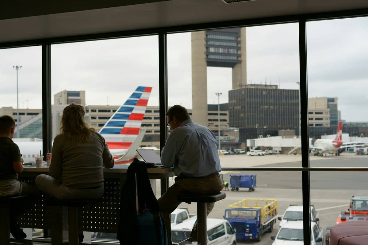 Passagiere sitzen an einem großen Fenster mit Blick auf das Rollfeld des Flughafens, ein Flugzeug der American Airlines und den Kontrollturm des Flughafens Boston Logan.
