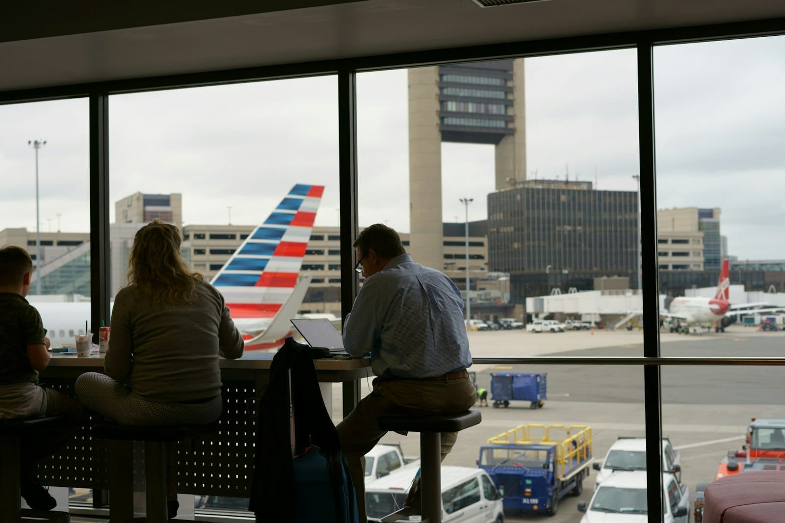 Viajantes sentados perto de grandes janelas dentro do terminal do Aeroporto Logan de Boston, com aviões e a torre de controle visíveis lá fora.