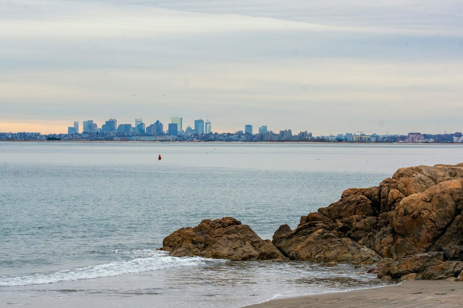 Vista de uma praia rochosa com o skyline de Boston ao fundo sob um céu nublado, com ondas suaves chegando à costa.
