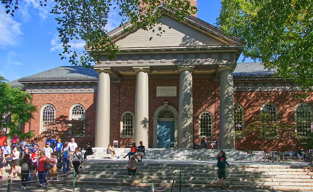 Un edificio clásico del campus de Harvard con columnas, fachada de ladrillo y estudiantes y visitantes reunidos en los escalones bajo el sol.