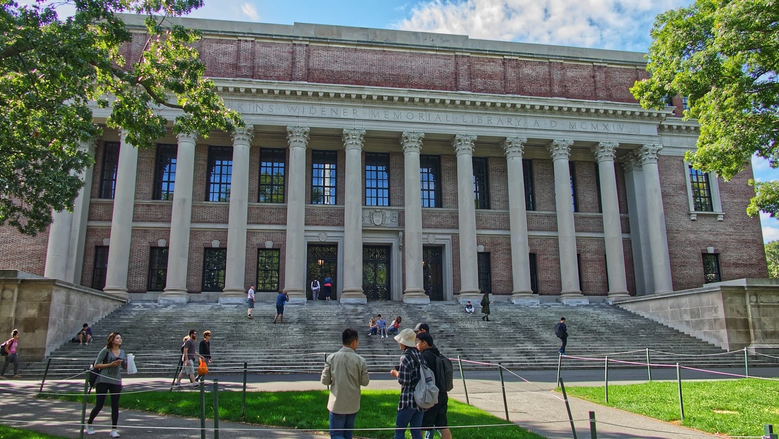 Vista frontal da icônica biblioteca da Universidade Harvard, com grandes colunas e visitantes nos degraus, emoldurada por árvores verdes sob a luz do dia.