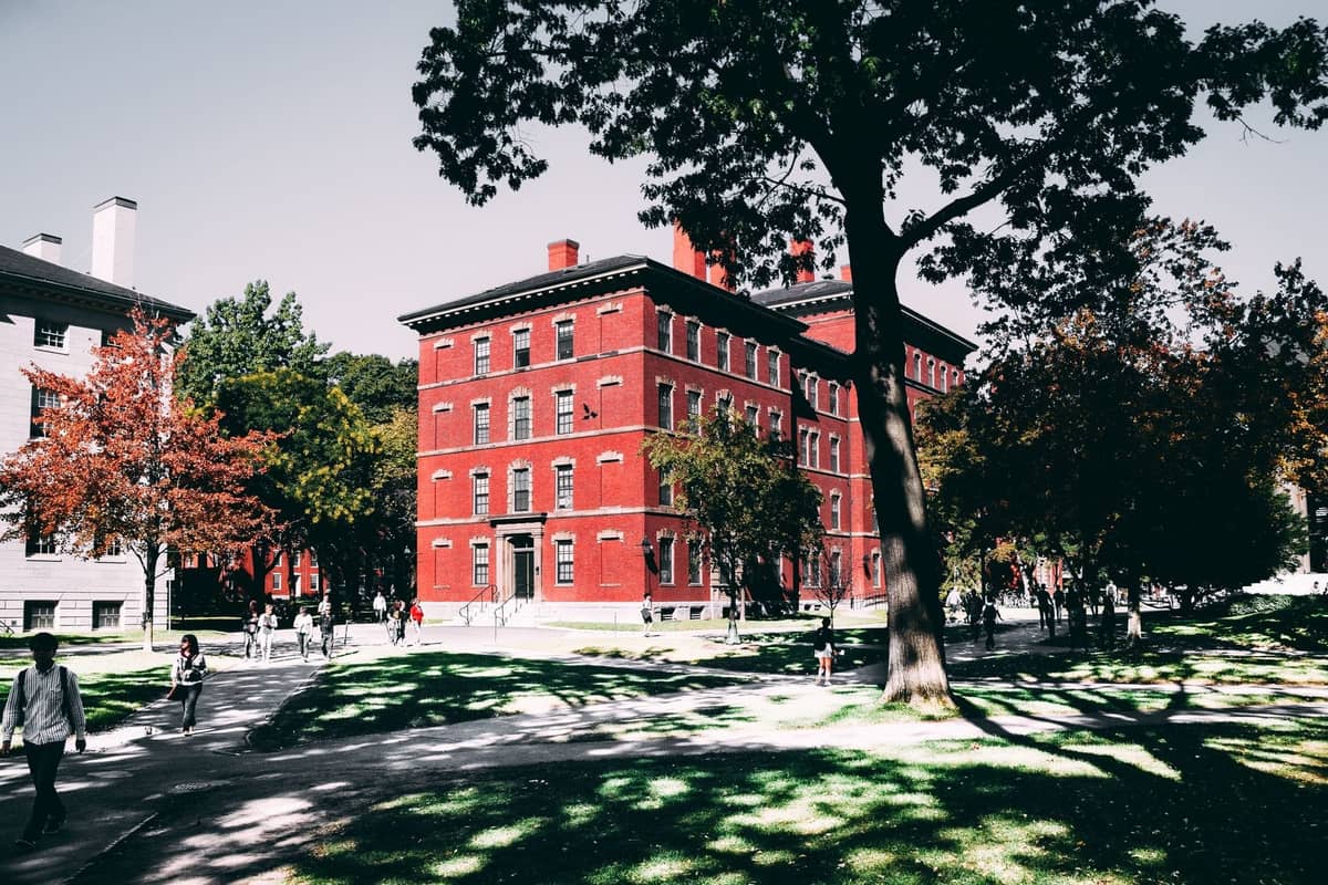 Personas caminando por el frondoso campus de Harvard con un clásico edificio de ladrillo rojo y árboles a finales de primavera o principios de otoño.