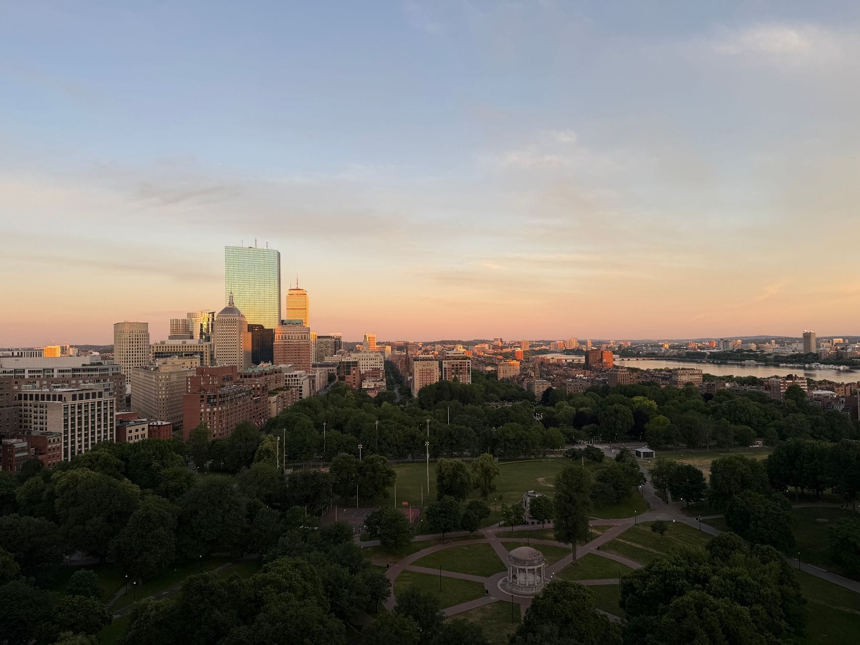 Una vista aerea del Boston Common al tramonto, con il verde del parco, i sentieri storici, lo skyline del centro e il fiume Charles sullo sfondo.
