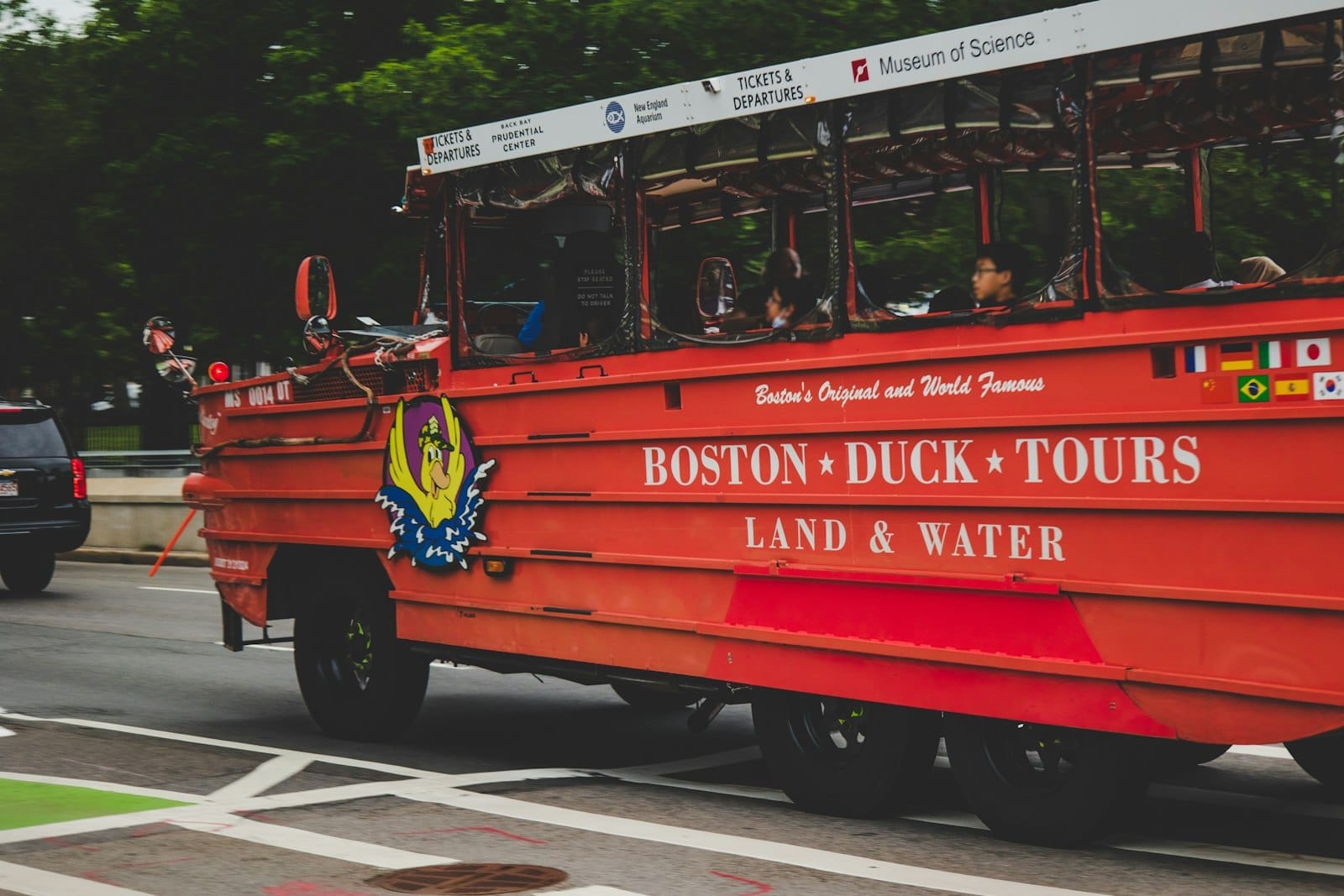 Veicolo anfibio rosso brillante dei Boston Duck Tours che percorre una strada cittadina con passeggeri e segnaletica del tour visibili, alberi sullo sfondo.