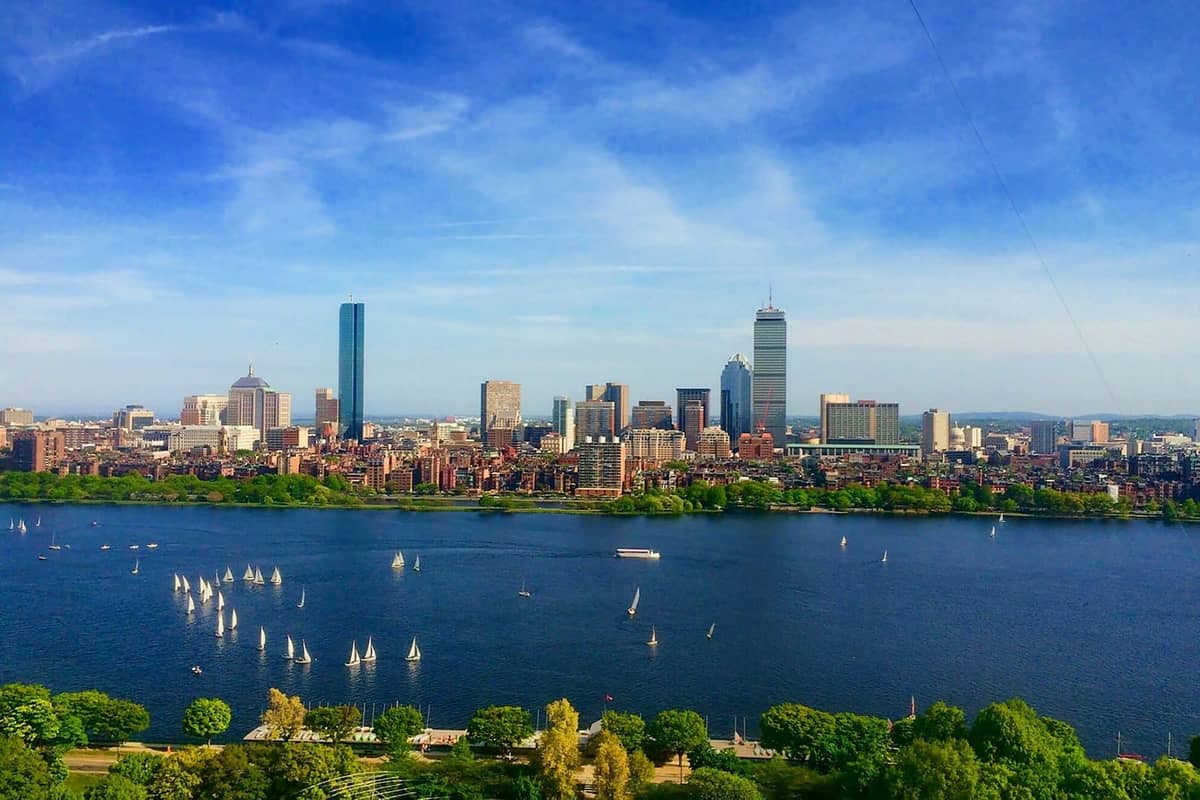 Blick auf die Skyline von Boston an einem hellen Sommertag über den Charles River, mit grünen Bäumen und weißen Segelbooten auf dem blauen Wasser.
