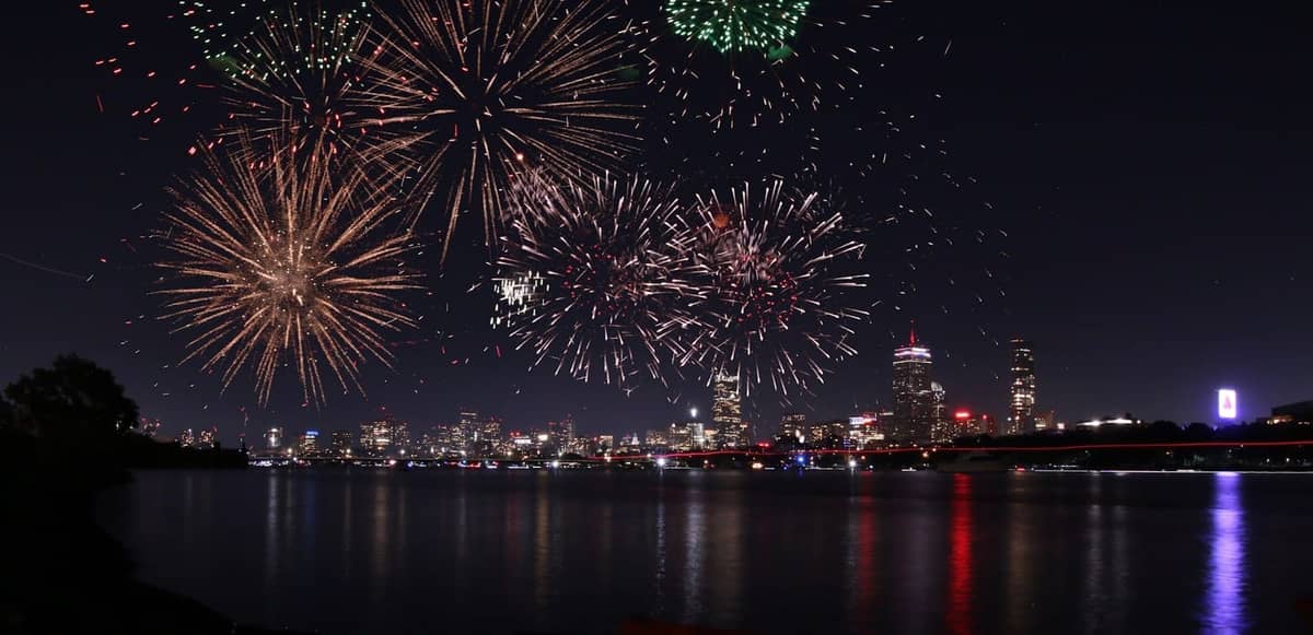 Nächtliche Ansicht der Skyline von Boston mit großem Feuerwerk zum 4. Juli über dem Charles River, Stadtlichter spiegeln sich im Wasser.