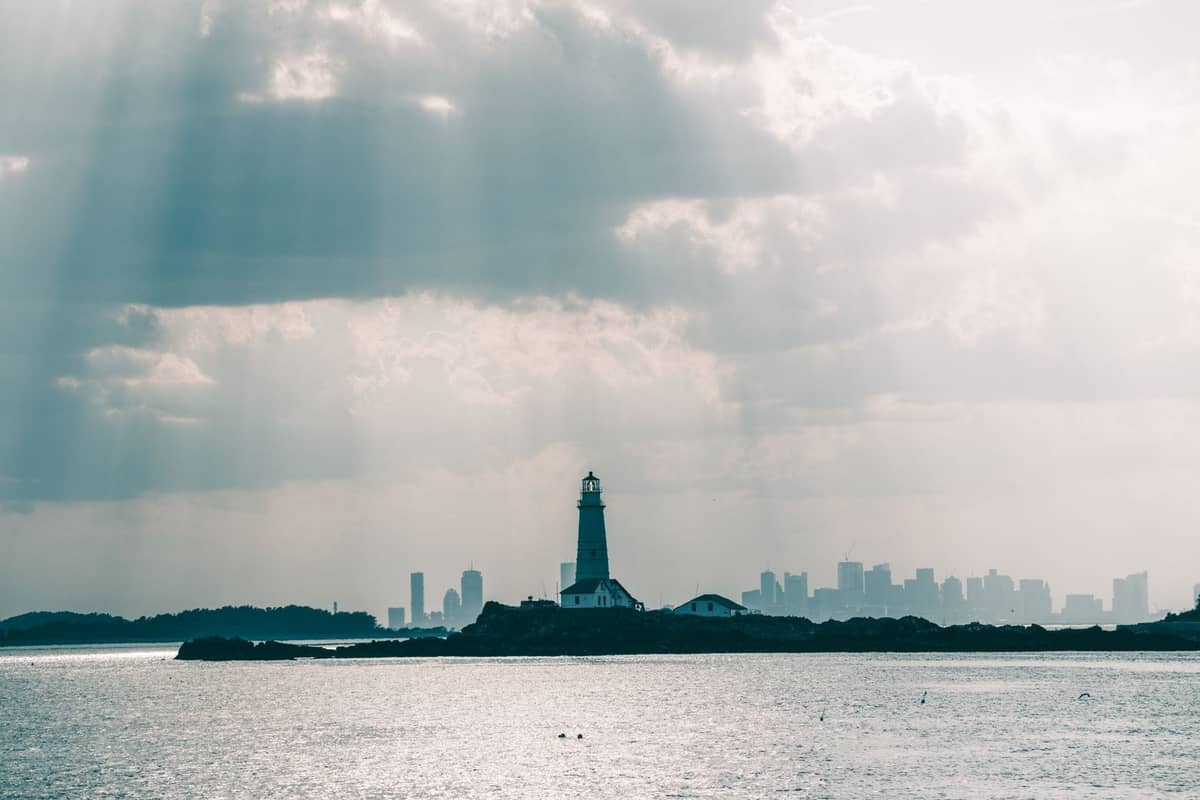 Faro en una isla en Boston Harbor con el horizonte de la ciudad visible al fondo y luz solar filtrándose entre las nubes.