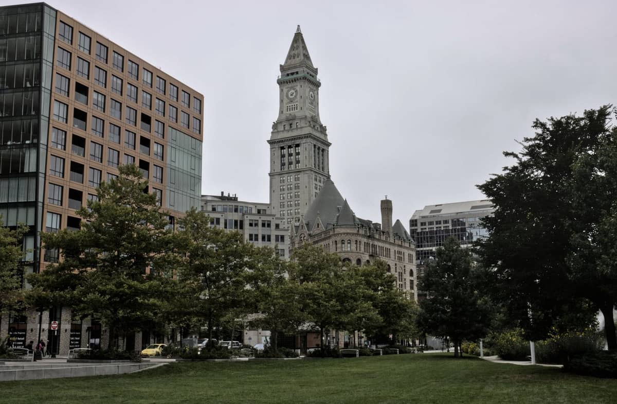 Boston'ın Custom House Tower'ı, bulutlu bir gökyüzünün altında şehir merkezinde yeşil park alanı ve modern binaların arkasında yükseliyor.
