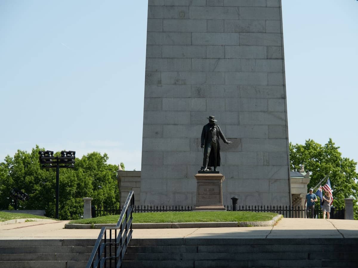 Güneşli bir günde Bunker Hill Monument'ın kaidesinde bir heykel ve Amerikan bayrağı tutan birkaç ziyaretçi.