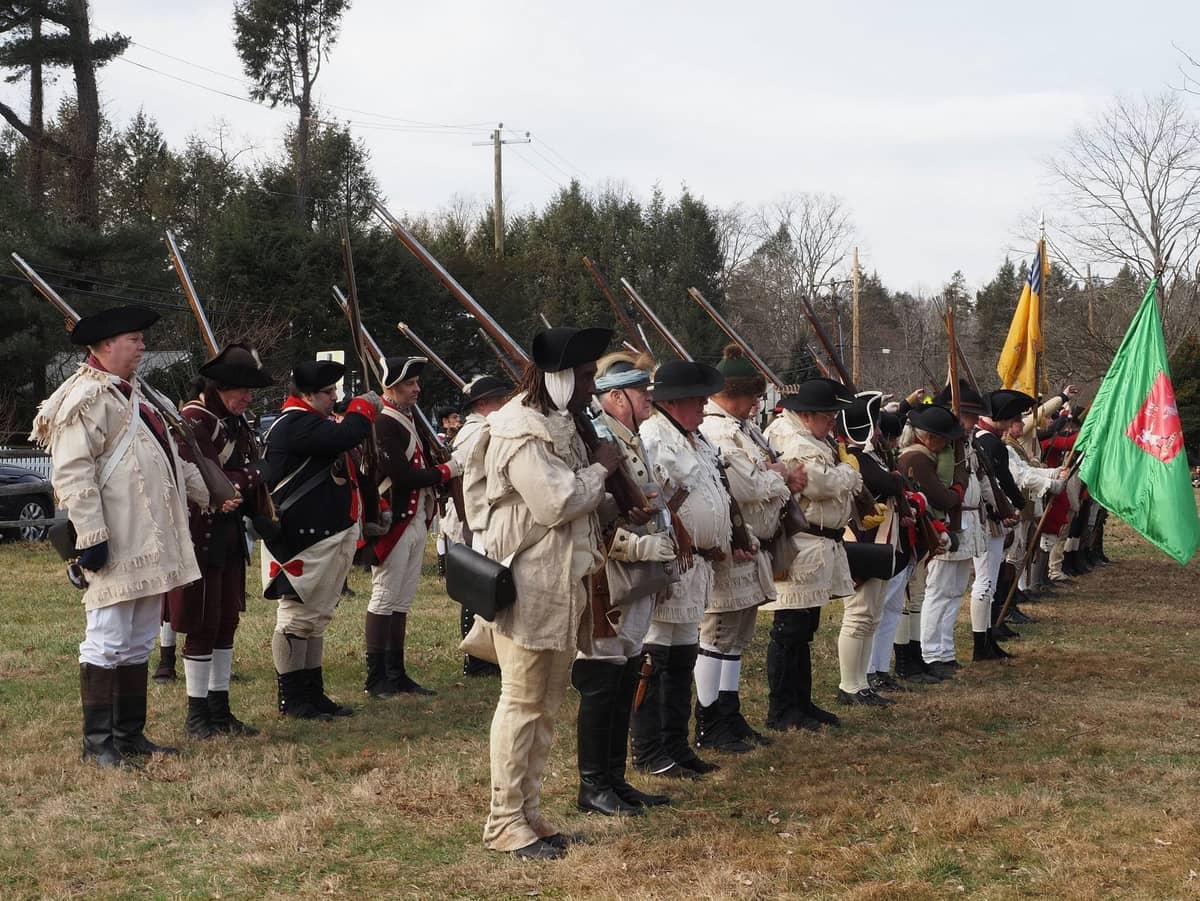 Çimenli bir alanda musketalarıyla duran kolonyal kıyafetler içindeki Devrim Savaşı yeniden canlandırmacıları grubu, Lexington veya Concord savaş alanını çağrıştırıyor.