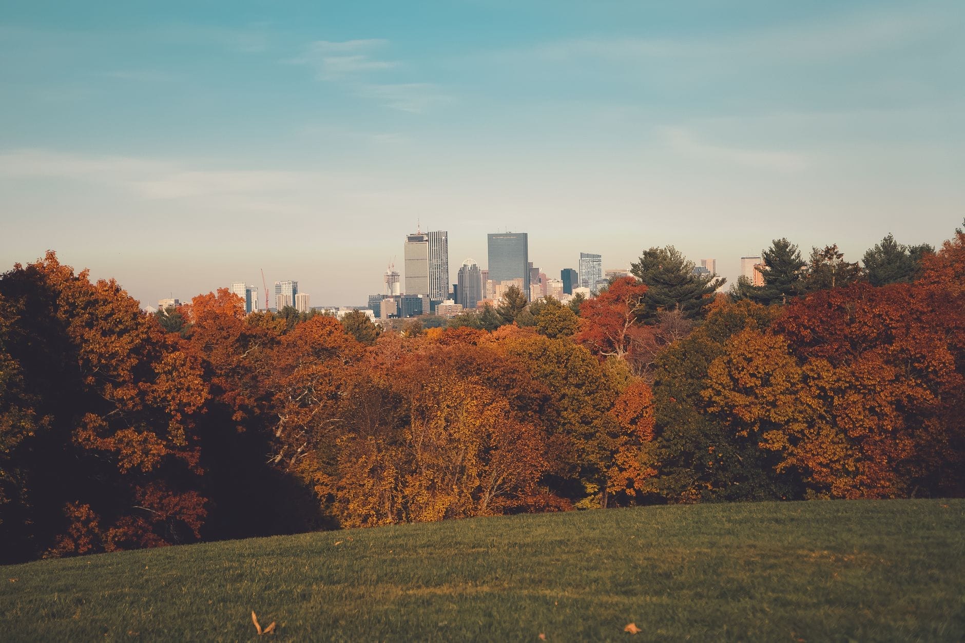 Skyline de Boston ao fundo, com uma explosão de folhagem outonal em tons de vermelho, laranja e dourado em primeiro plano, sob um céu limpo.