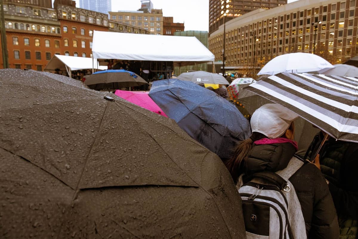 Menschenmenge mit Regenschirmen bei einer Veranstaltung im Freien auf einem Stadtplatz in Boston mit erkennbaren Gebäuden der Innenstadt im Hintergrund.