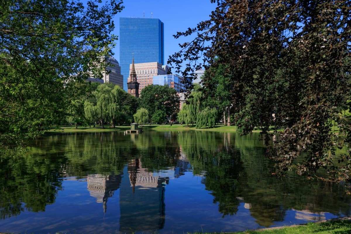 Boston Public Garden im Sommer mit einem spiegelnden Teich, üppigen grünen Bäumen und der Skyline der Stadt im Hintergrund unter einem klaren blauen Himmel.