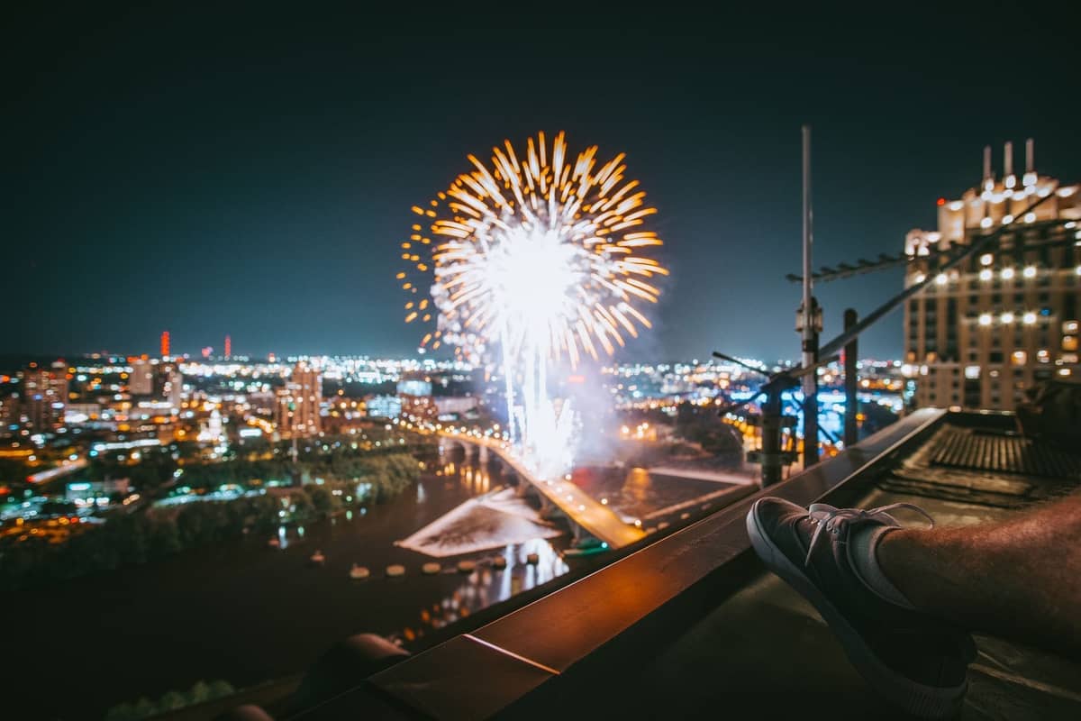Nächtliche Aussicht auf Feuerwerk über dem Fluss mit beleuchteter Stadtsilhouette und Brücke, von einem Dach in Boston aus gesehen.