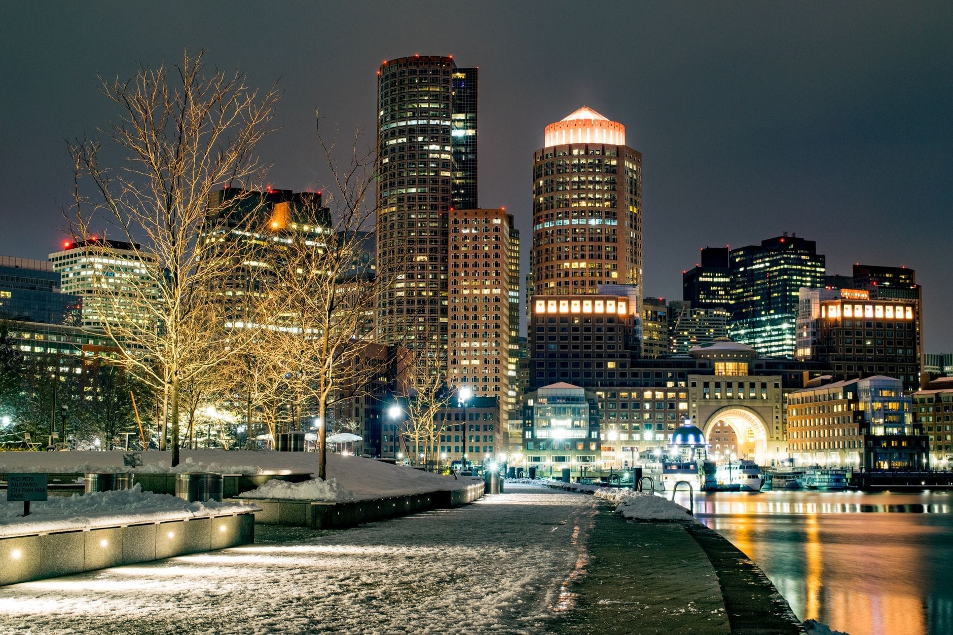 Skyline do centro de Boston brilhando à noite com neve no chão, luzes da cidade refletidas na água e uma atmosfera tipicamente hibernal.
