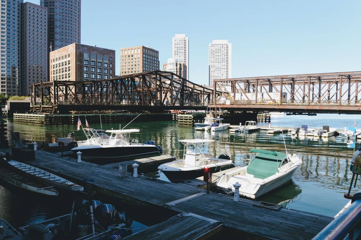 Escena del paseo marítimo de Boston con lujosos barcos atracados en una marina, edificios del horizonte urbano y un puente histórico al fondo.