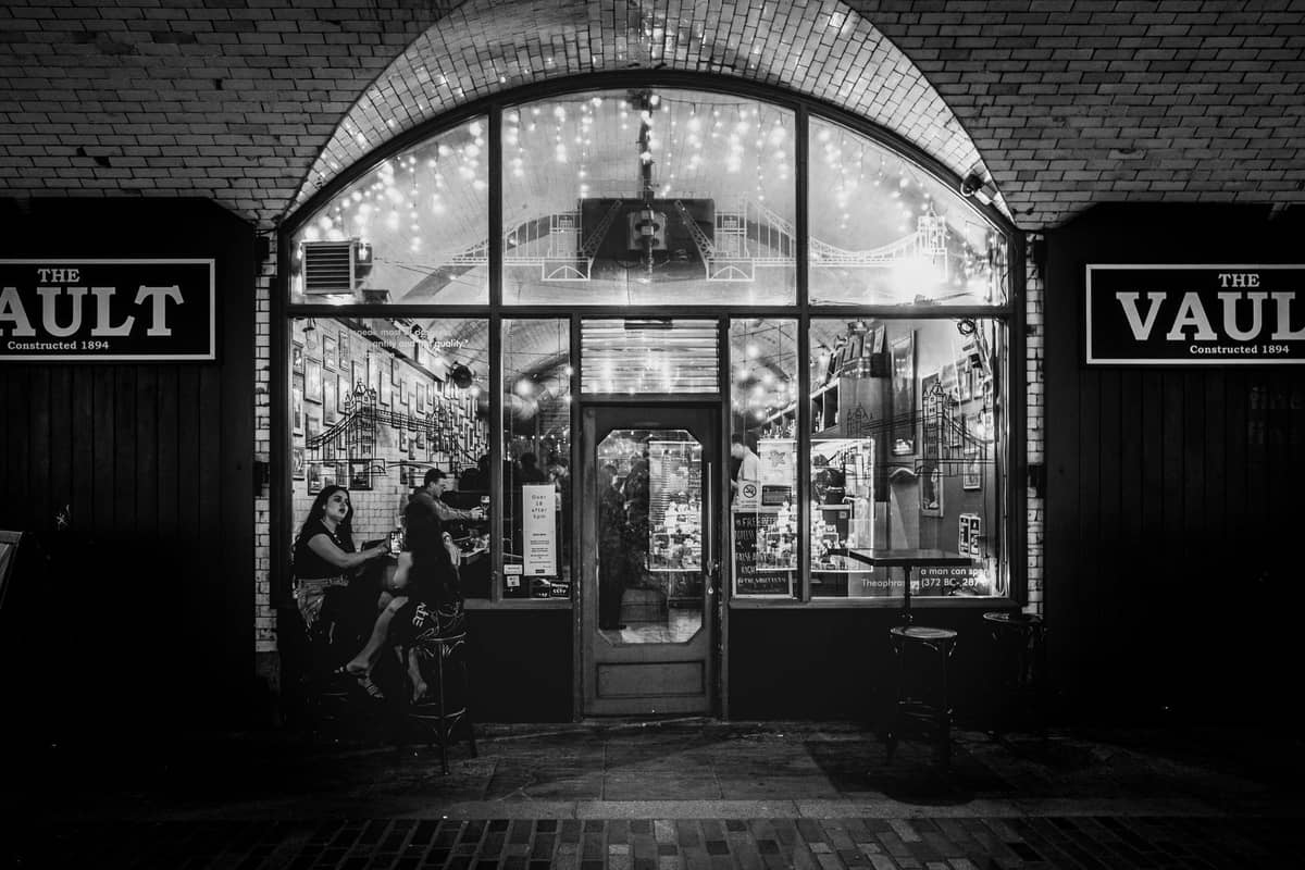 Foto in bianco e nero dell'ingresso del bar The Vault a Boston di notte, con persone sedute fuori sotto luci decorative.
