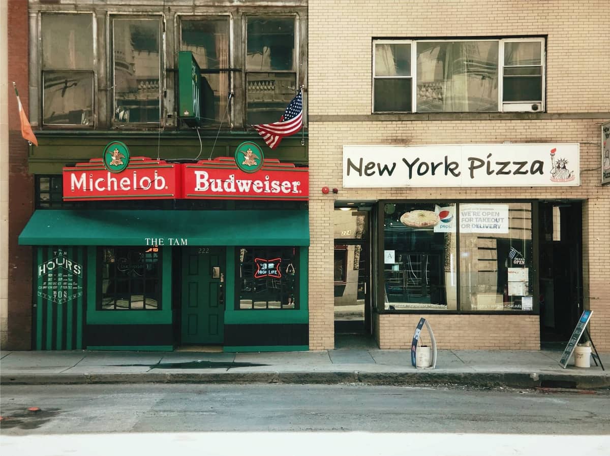 Vetrine del bar The Tam e di New York Pizza su una strada cittadina, con insegne verdi e rosse e un'atmosfera classica di quartiere.