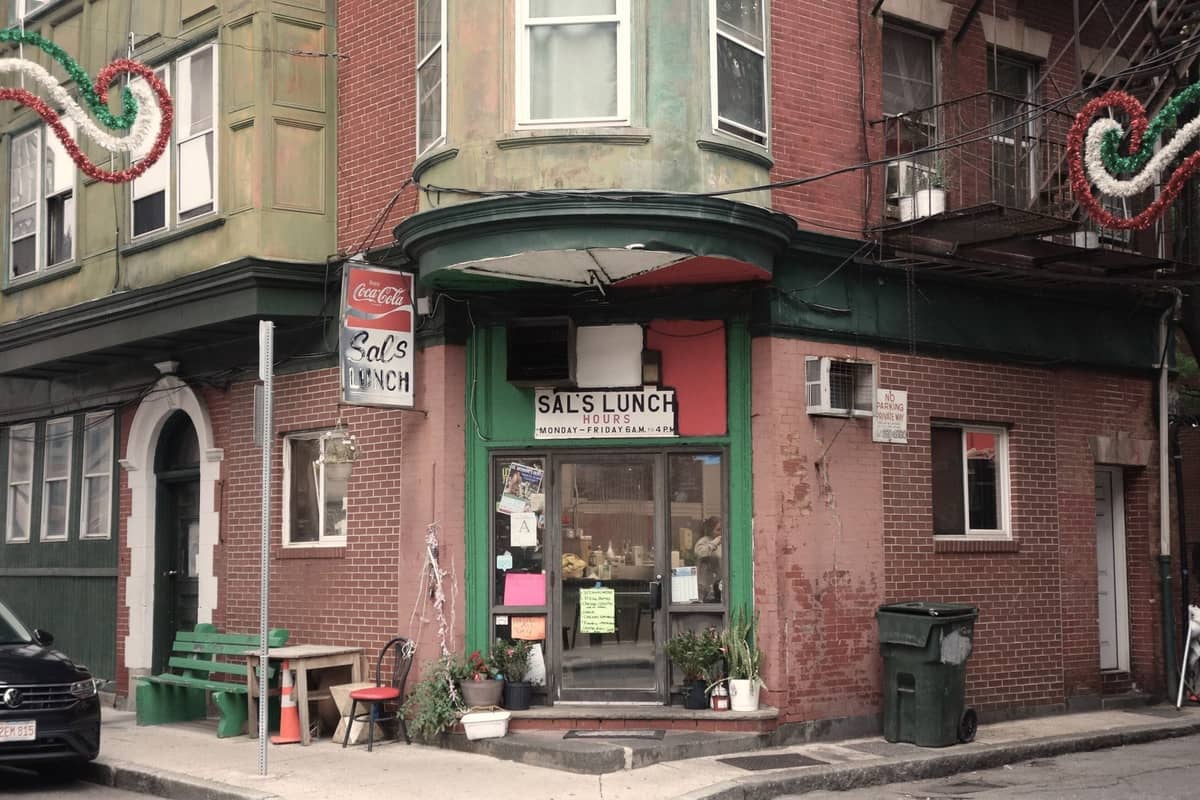 Straßenansicht von Sal's Lunch, einem bescheidenen Ecklokal im North End von Boston, mit Schildern, Bänken und Pflanzen draußen.