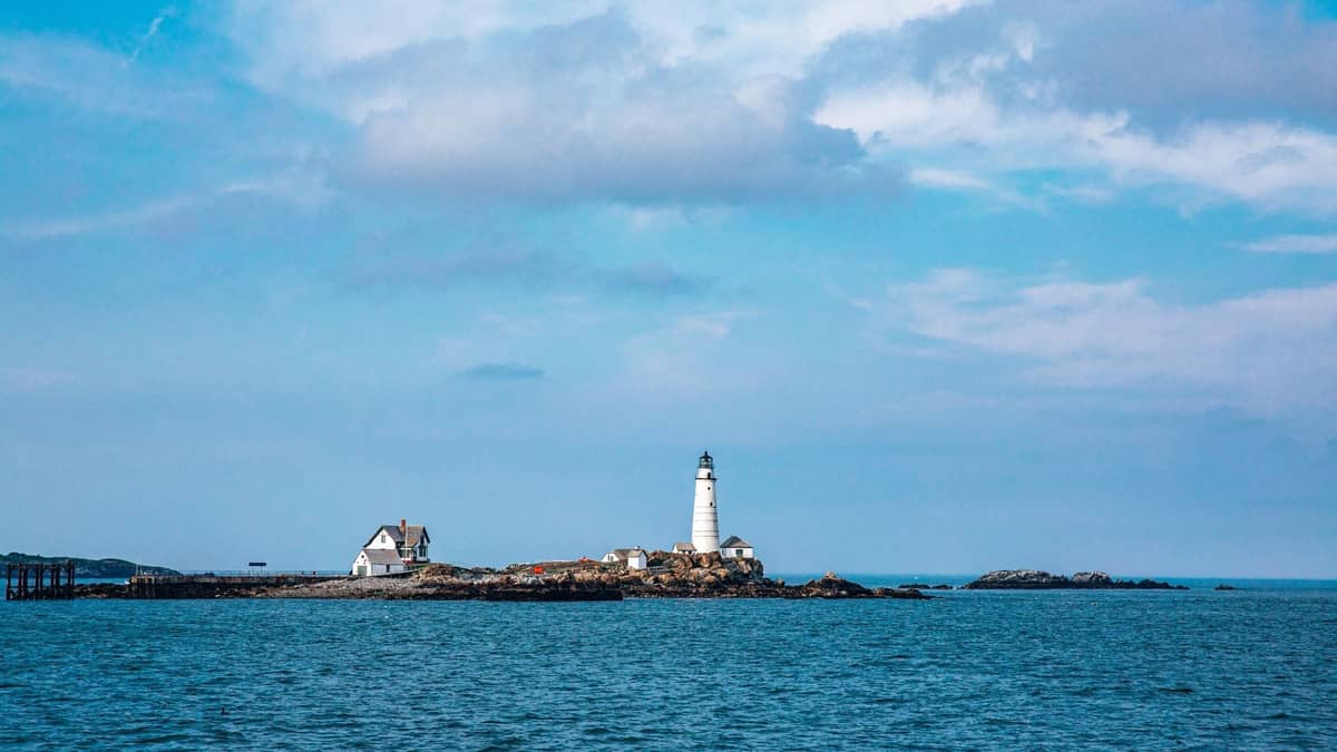 Mała wyspa w Boston Harbor z latarnią morską, domem i skalistym brzegiem, otoczona błękitną wodą pod częściowo zachmurzonym niebem.