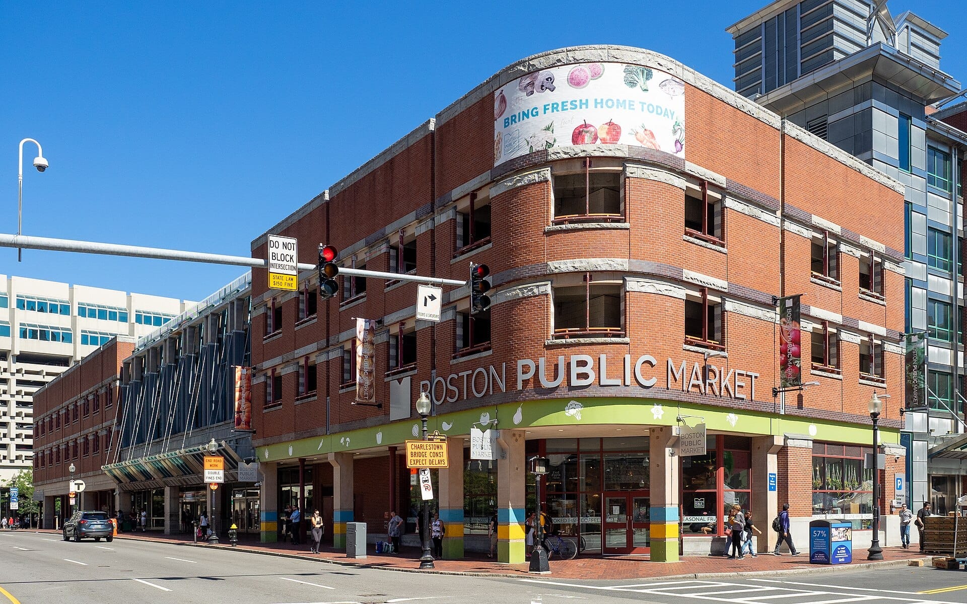 Edificio in mattoni del Boston Public Market con grandi finestre e insegne colorate in una giornata di sole, situato in una trafficata strada cittadina.