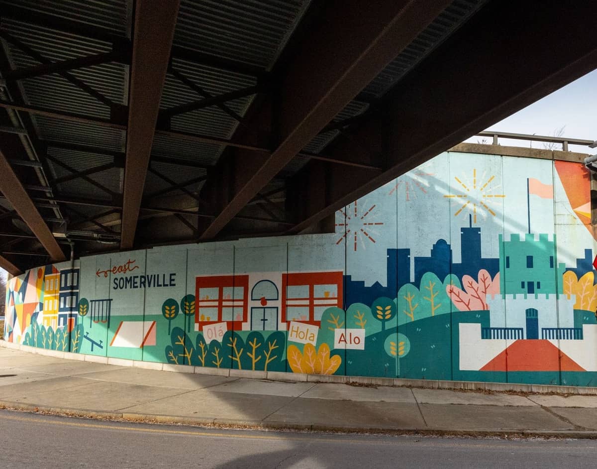 Une fresque colorée avec l'inscription 'greetings from Somerville' sous un pont, avec la skyline de Boston et des icônes locales illustrées.