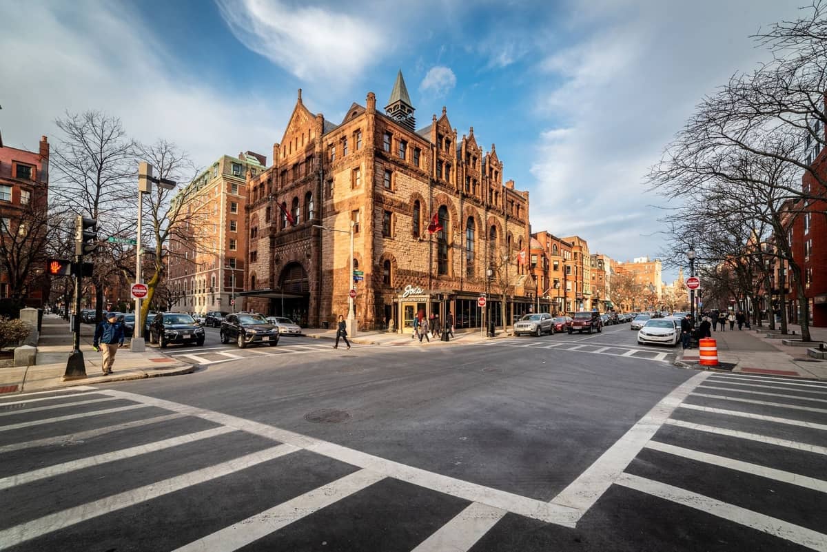 Vue large de l'intersection de Newbury Street à Boston avec ses bâtiments historiques en grès brun, ses devantures de magasins et ses piétons sous un ciel bleu.