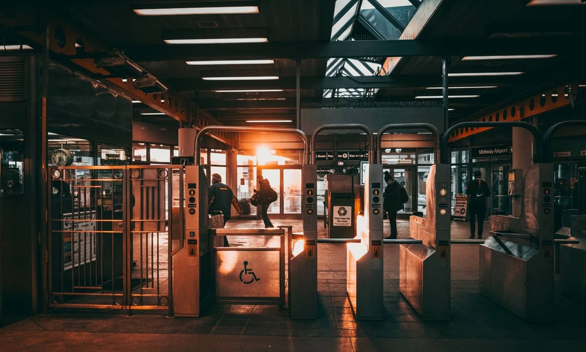 Pasażerowie przechodzący przez bramki biletowe na nowoczesnej stacji tranzytowej z promieniami słońca w tle.
