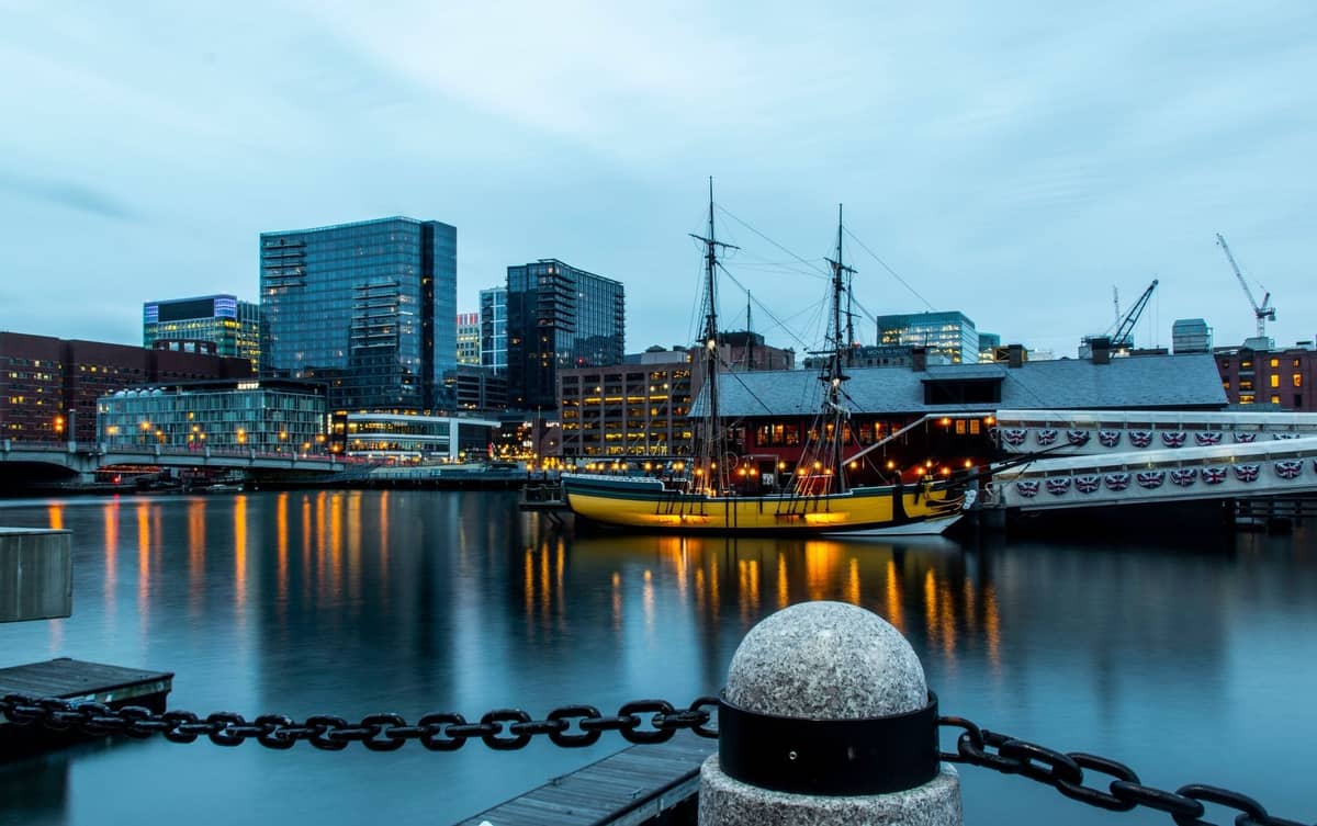 Evening view of the Boston Tea Party Ships & Museum with a replica 18th-century ship docked by the waterfront and city buildings in the background.