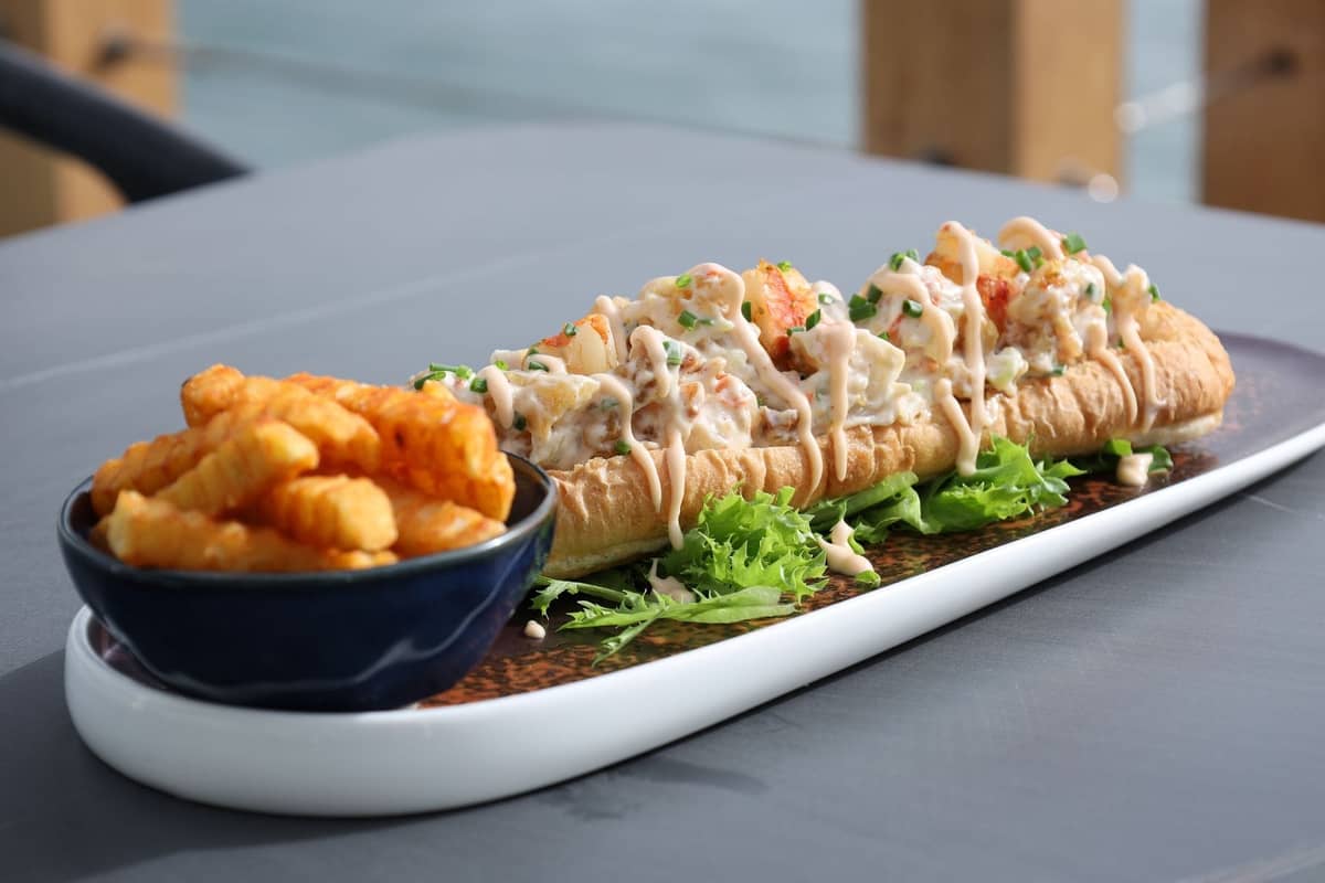 Un lobster roll nappé de sauce est servi dans une longue assiette avec des légumes verts et des frites gaufrées, sur une table en bord de mer.