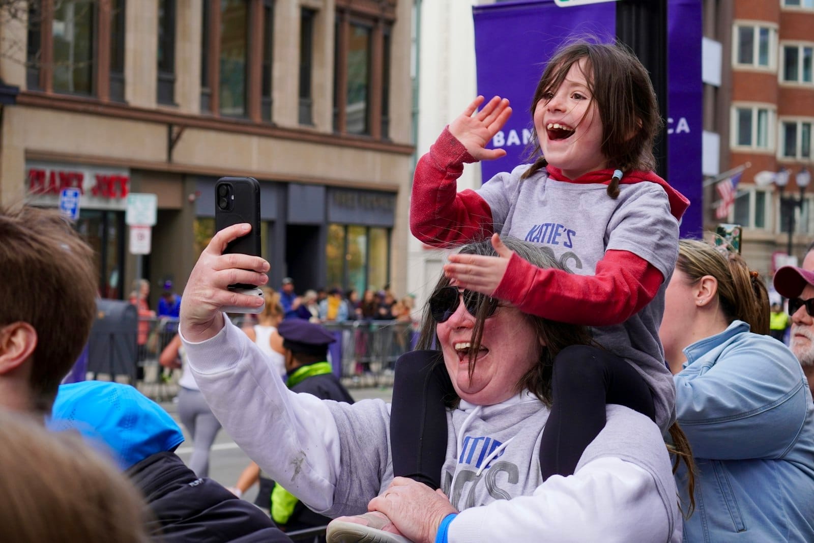 Criança sorridente nos ombros de um adulto acena durante um evento animado em uma ensolarada rua de Boston ladeada por edifícios históricos.