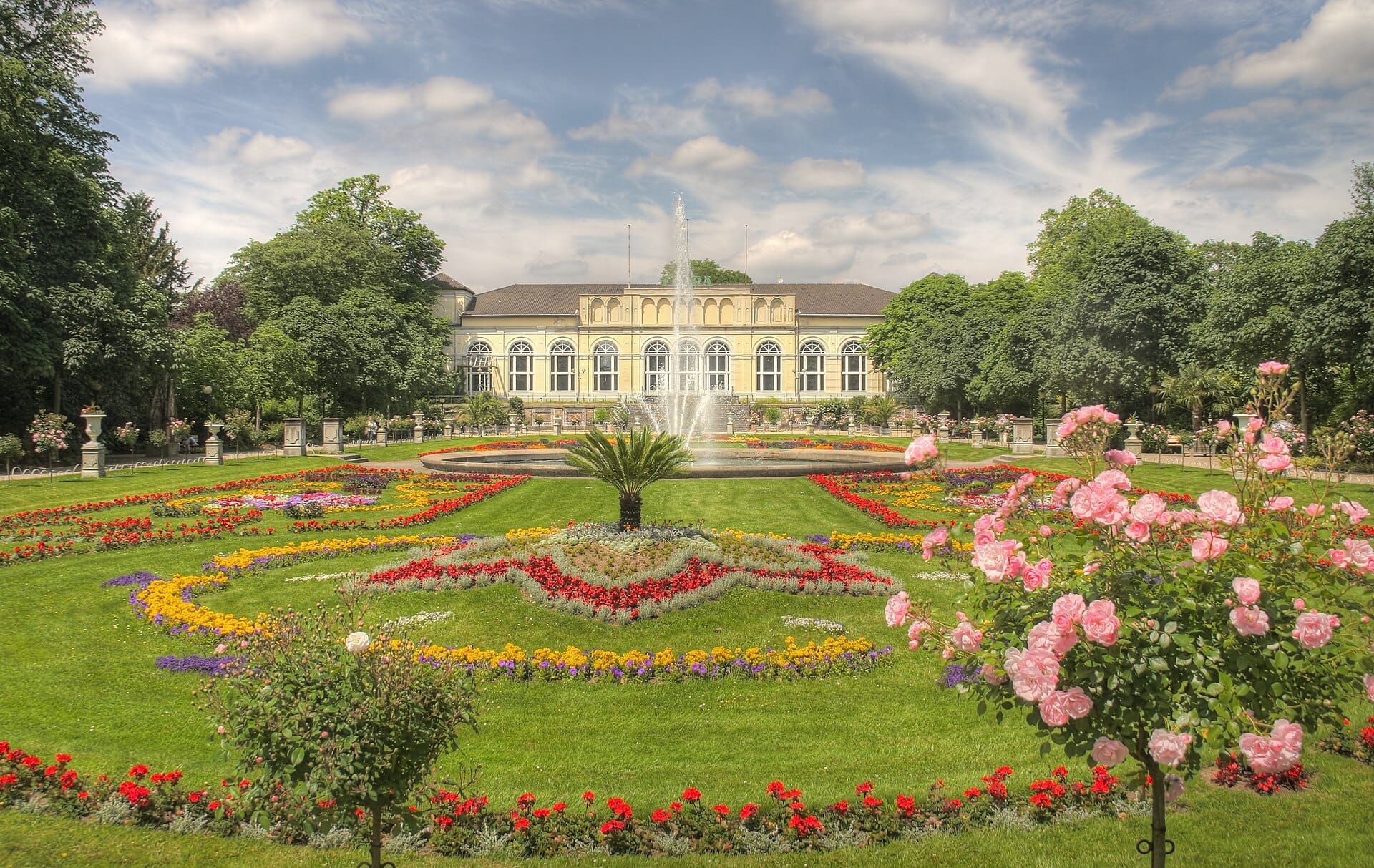 แปลงดอกไม้สมมาตรและน้ำพุกลางอยู่ด้านหน้าอาคาร Flora อันเก่าแก่ใน Flora und Botanischer Garten Köln ในวันที่แดดสดใส