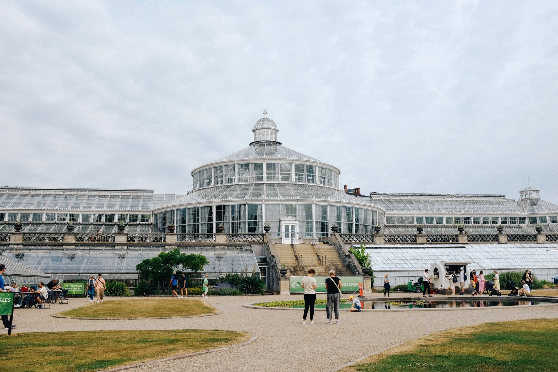 Veduta ampia dell'iconica serra vittoriana del Giardino Botanico di Copenaghen, con persone che passeggiano e si rilassano sui sentieri circondati dal verde sotto un cielo aperto.