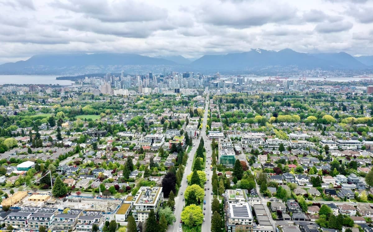 Luftaufnahme der grünen Stadtteile von Vancouver und der Skyline der Stadt mit üppig baumgesäumten Straßen und fernen Bergen an einem bewölkten Tag.