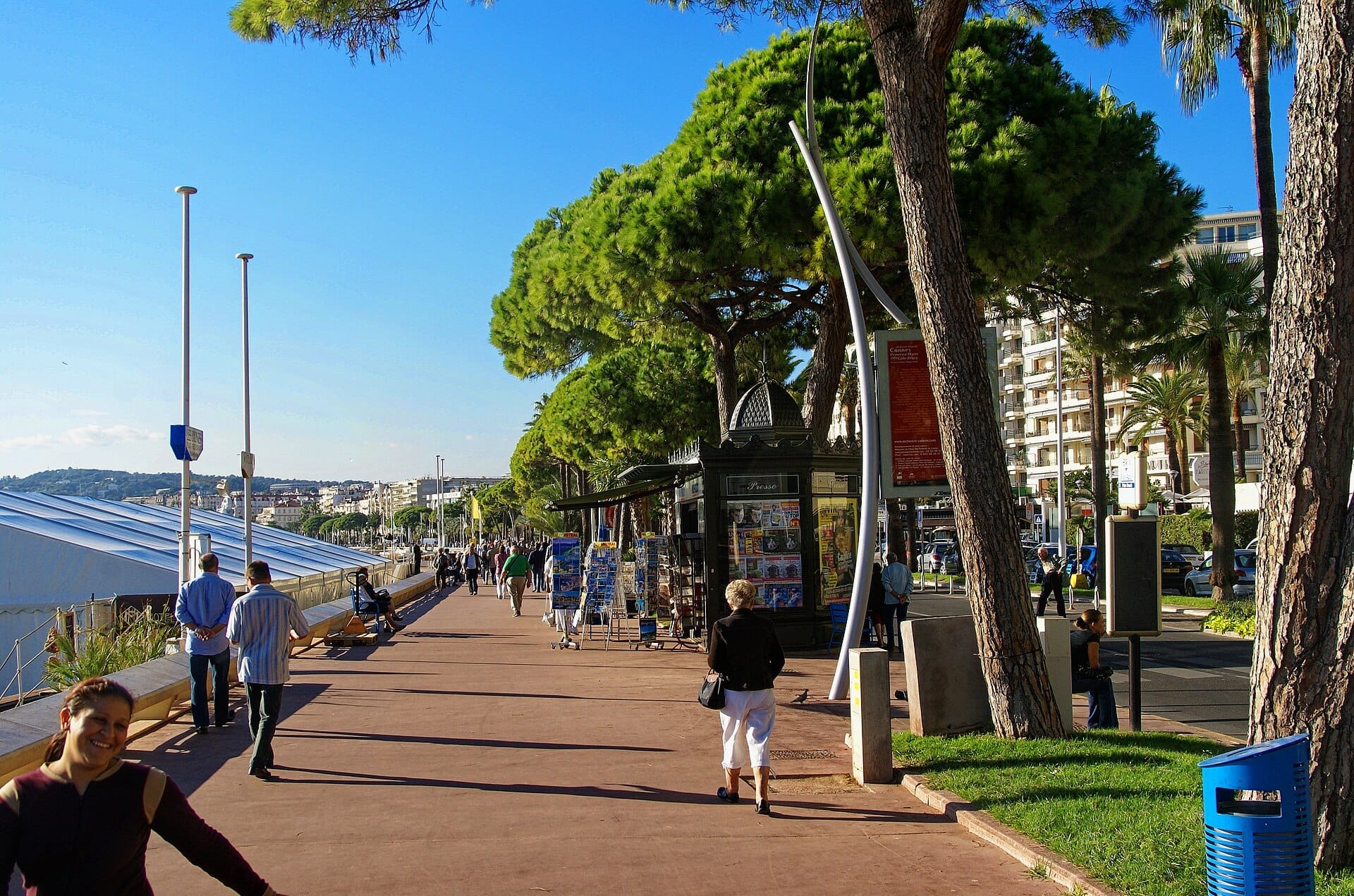Le persone passeggiano lungo il Boulevard de la Croisette fiancheggiato da palme a Cannes, con edicole e edifici moderni sotto un cielo azzurro limpido.