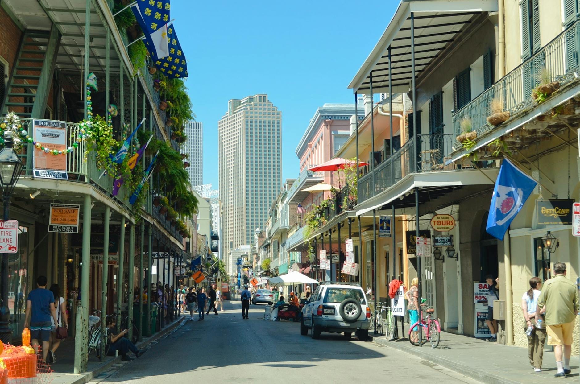 Una vista diurna e luminosa di Bourbon Street a New Orleans con bandiere colorate, balconi in ferro battuto, piante, passanti e grattacieli sullo sfondo sotto un cielo azzurro.