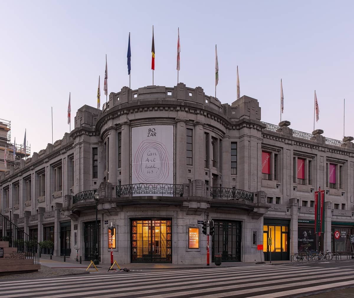 Fachada Art Déco do BOZAR Centre for Fine Arts em Bruxelas, com bandeiras hasteadas no topo e luzes quentes brilhando na entrada principal.