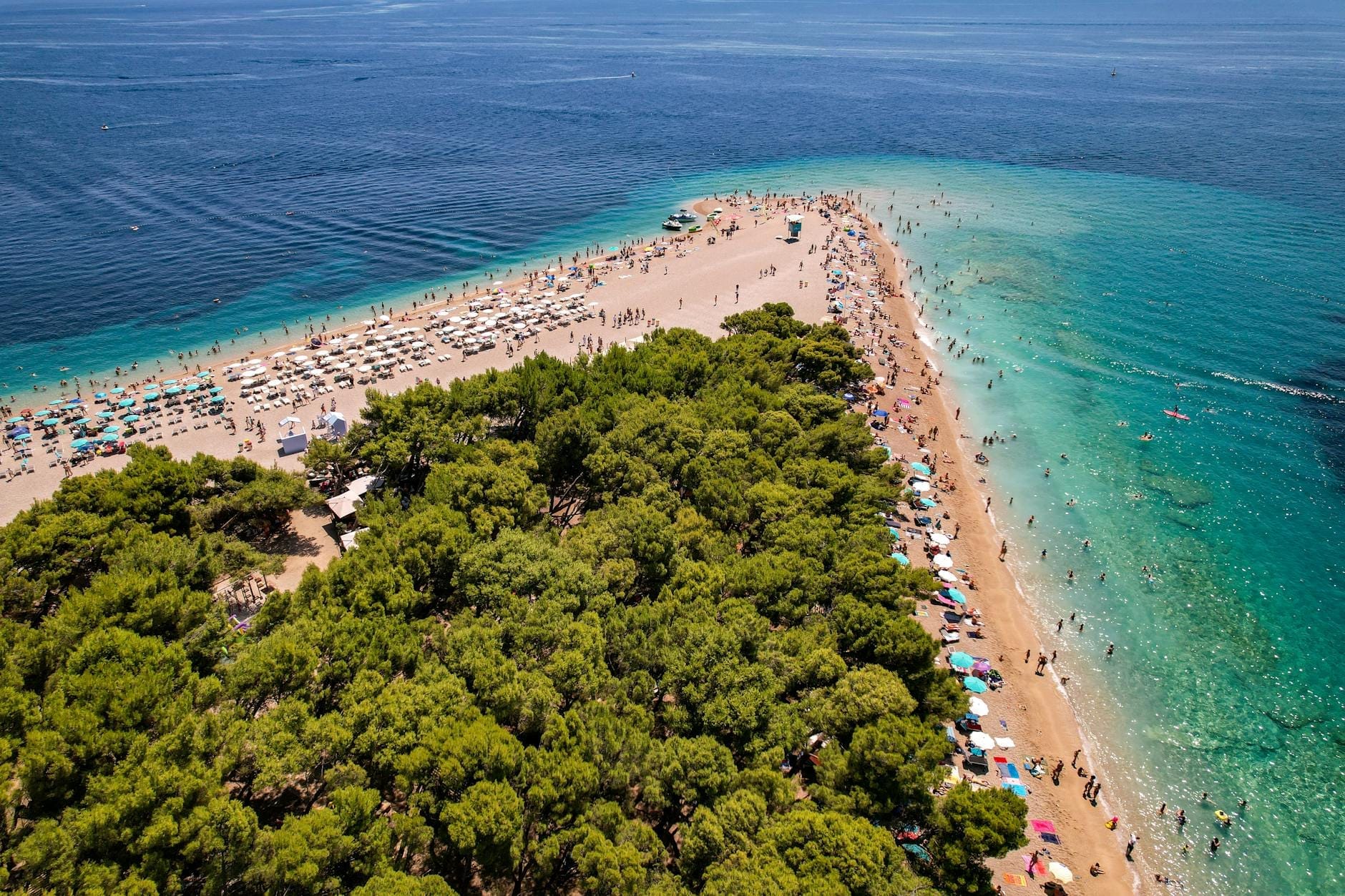 มุมสูงของหาด Zlatni Rat อันโด่งดังบนเกาะบรัช โครเอเชีย น้ำทะเลสีฟ้าใส คนเล่นน้ำ ร่มชายหาดและป่าสนเขียวชอุ่ม