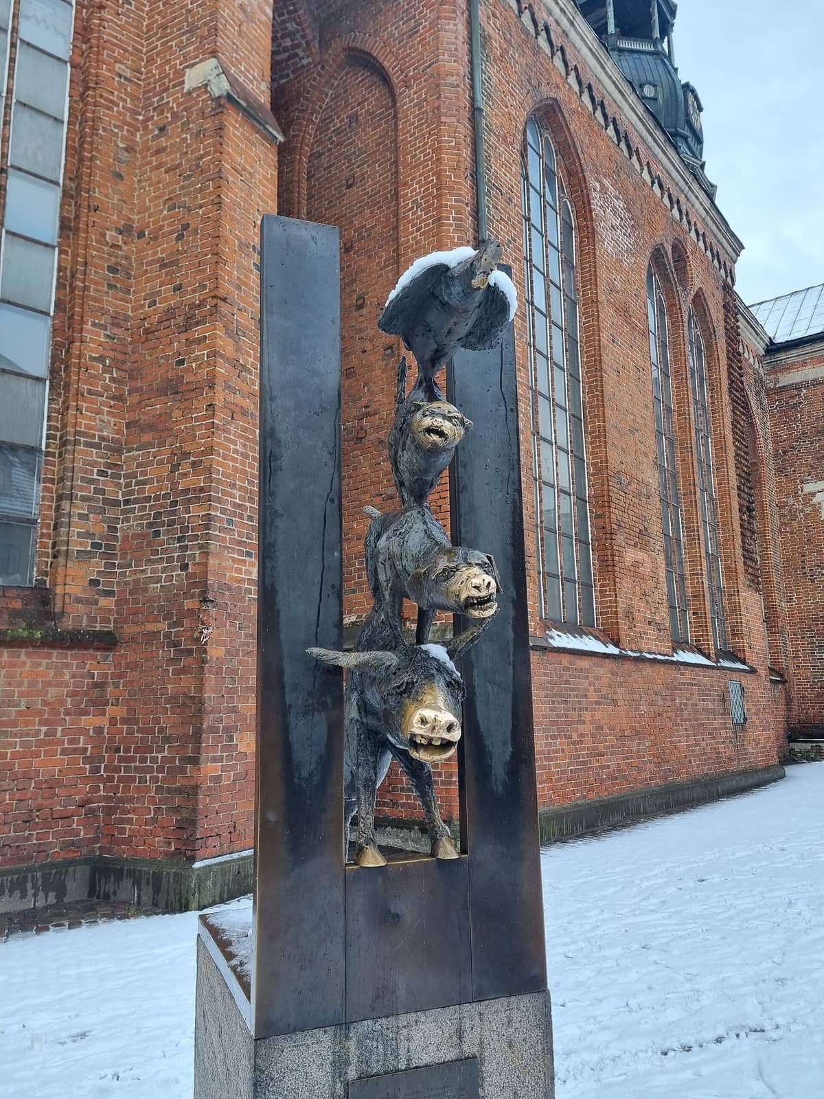 Bronzestatue der Bremer Stadtmusikanten mit Schnee auf den Tieren, vor Rigas roter mittelalterlicher Backsteinkirche und Winterlandschaft.