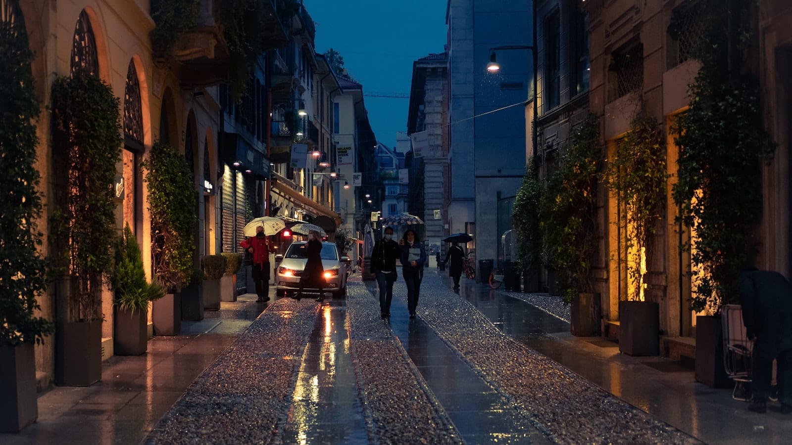 บรรยากาศยามเย็นของถนนหินกรวดในย่าน Brera มิลาน รายล้อมด้วยร้านบูติกและร้านอาหาร แสงไฟอบอุ่น มีผู้คนเดินถือร่ม