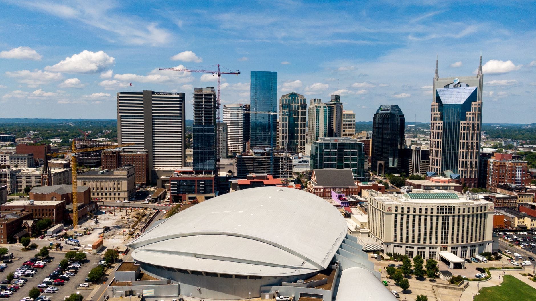 Vista aérea del Bridgestone Arena con su techo curvo en el centro de Nashville, rodeado de modernos rascacielos y grúas de construcción bajo un cielo azul.