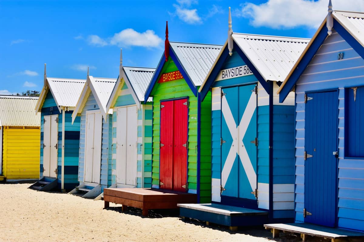 A row of brightly painted Brighton Beach Bathing Boxes on the sand at Dendy Street Beach, Melbourne, under a blue sky.