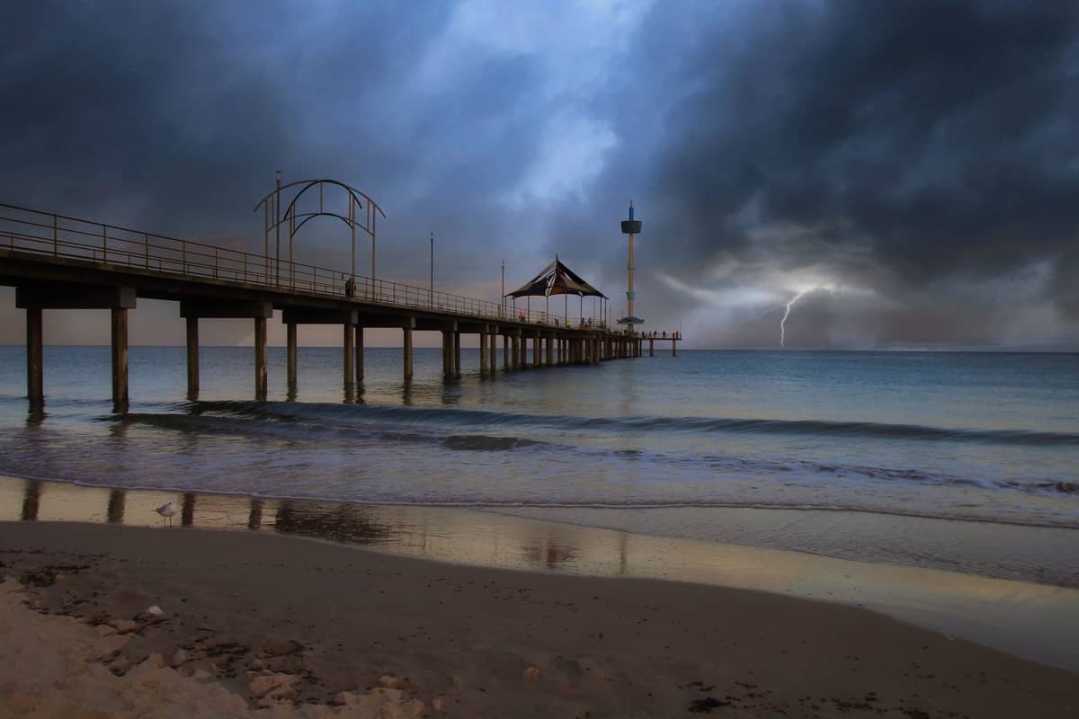 Dramatyczny wieczorny widok na molo w Brighton wychodzące w spokojne morze pod burzowym niebem, z błyskawicą w tle i łagodnymi falami na plaży.