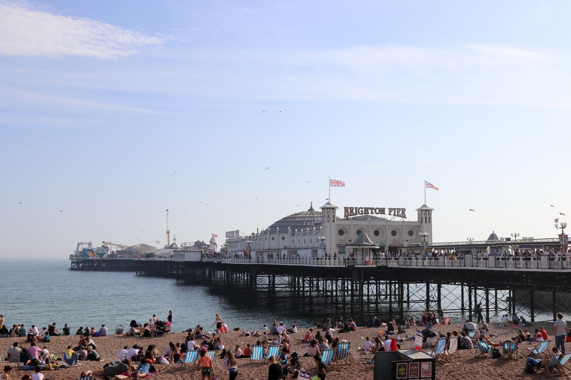 Brighton Pier extending into the sea with people relaxing on the lively pebble beach under a clear blue sky.