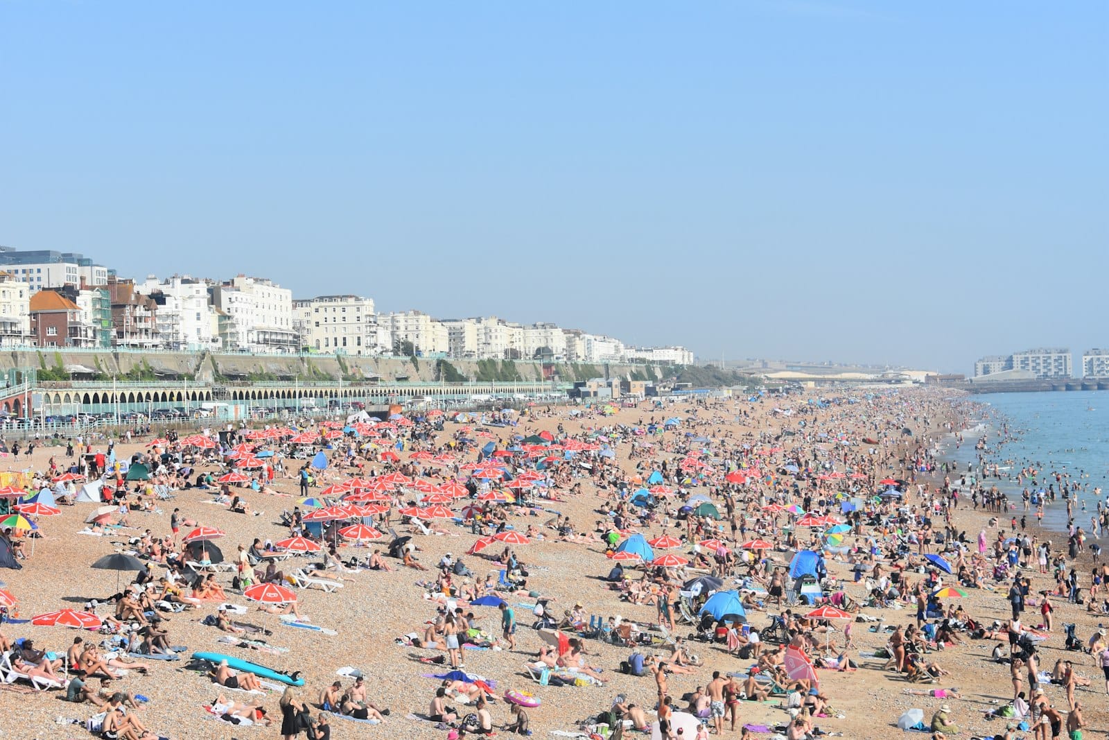 Die Strandpromenade von Brighton an einem sonnigen Tag mit einem belebten Kieselstrand, sonnenbadenden Menschen, bunten Sonnenschirmen und Gebäuden an der Strandpromenade im Hintergrund.