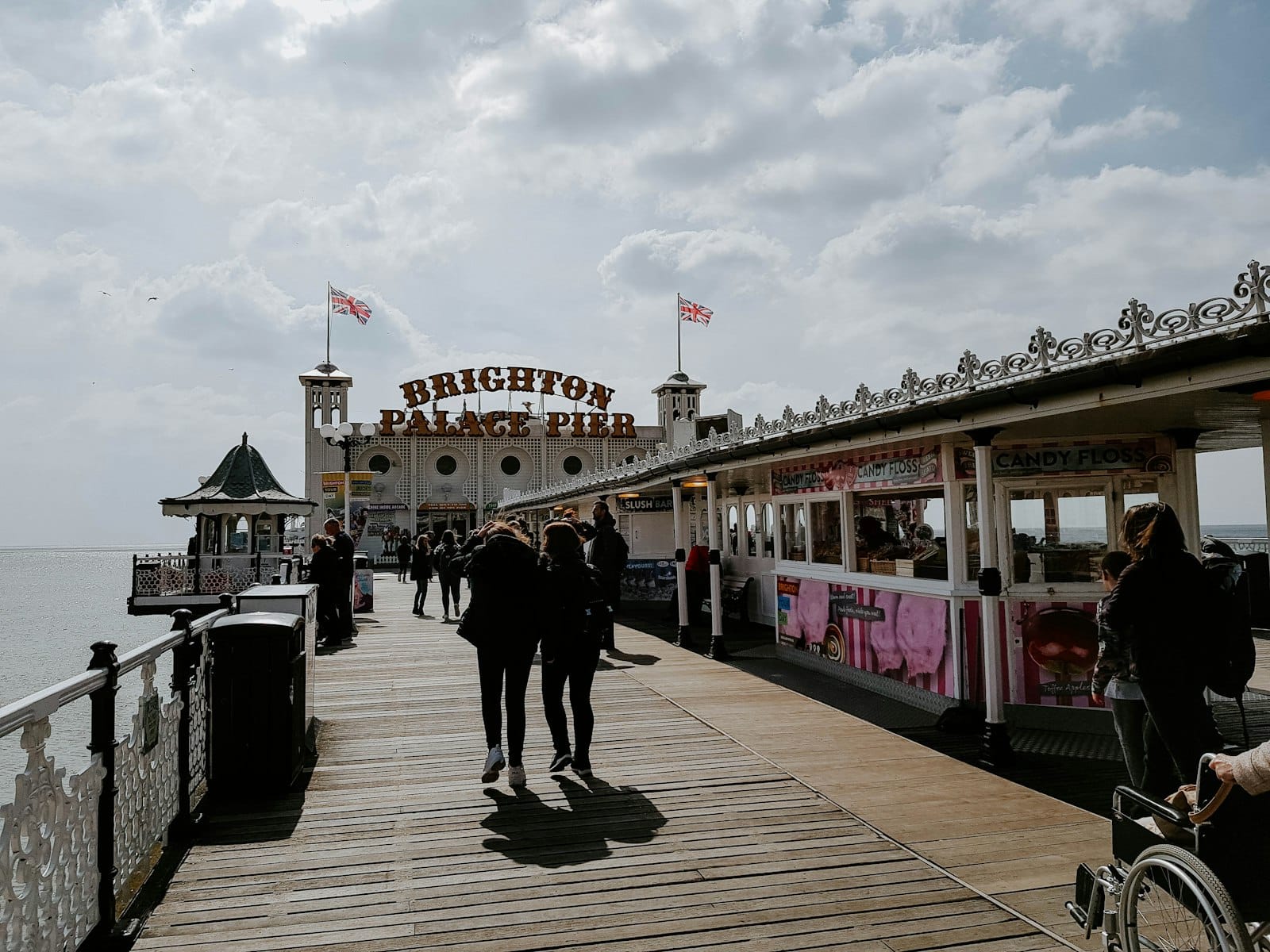 Menschen, die an einem teilweise bewölkten Tag auf dem Brighton Palace Pier spazieren, mit Vergnügungsständen und dem Piereingang im Bild.